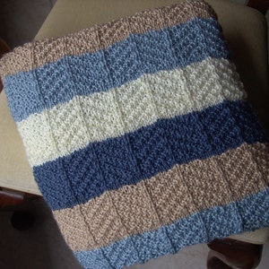 May include: A striped knitted throw blanket in shades of beige, blue, and white. The blanket is made with a textured stitch pattern and has a soft, cozy feel.