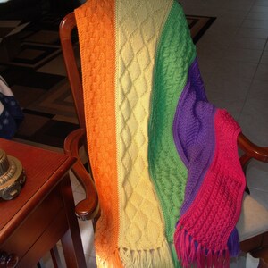 May include: A rainbow colored crocheted throw blanket with fringe on the ends. The blanket is draped over a wooden chair.