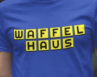 May include: A royal blue short-sleeved t-shirt featuring the text WAFFEL HAUS printed in black letters inside individual yellow square boxes arranged in two rows across the center of the chest area of the garment.