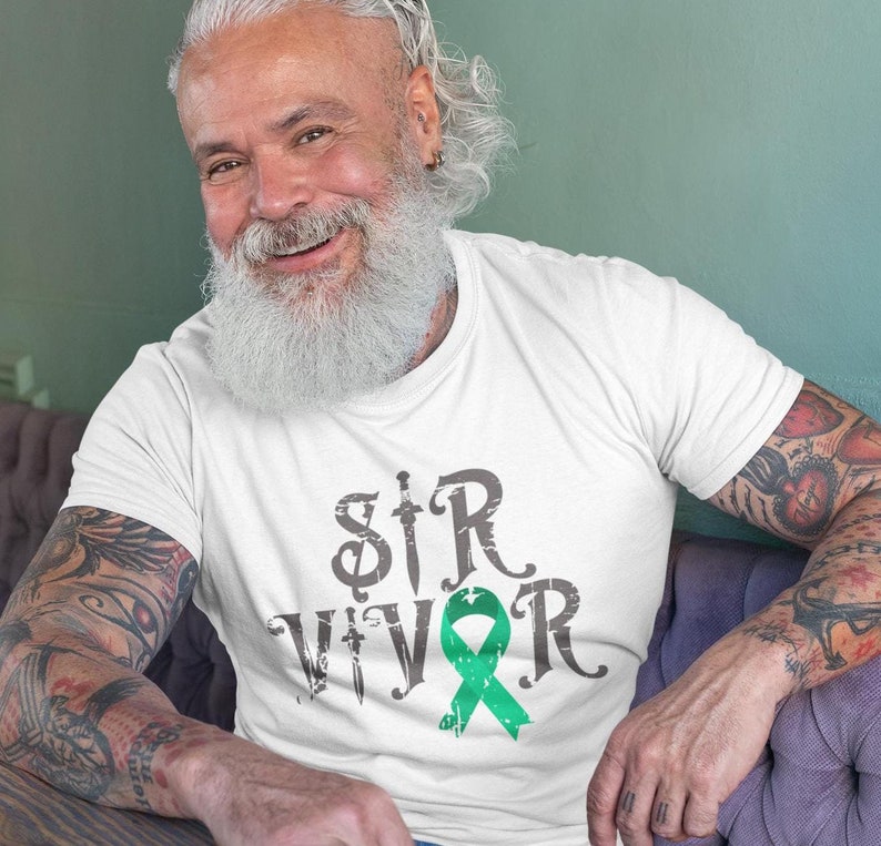Puede incluir: Camiseta blanca con la palabra "SURVIVOR" en letras grises desgastadas y una cinta verde. La letra "S" se reemplaza con una espada. La imagen muestra a una persona usando la camiseta.