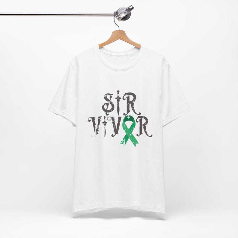 Puede incluir: Camiseta blanca con la palabra "SURVIVOR" en fuente negra desgastada y un lazo verde. La camiseta est&aacute; colgada de una percha de madera.
