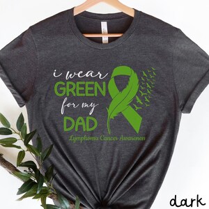 Könnte beinhalten: Dunkelgraues T-Shirt mit Melange-Effekt mit einem grünen Band und einem Federdesign. Der Text "Ich trage Grün für meinen Vater" und "Lymphom-Krebs-Bewusstsein" ist auf das T-Shirt gedruckt.