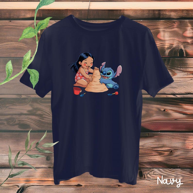 May include: Navy blue t-shirt featuring a graphic of Lilo and Stitch building a sandcastle. Lilo is wearing a red floral dress, and Stitch is blue. The shirt is hanging on a wooden background.