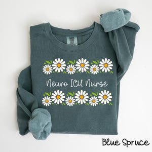 Pode incluir: Uma camisa de frio azul com uma borda de margaridas brancas e o texto "Neuro ICU Nurse".
