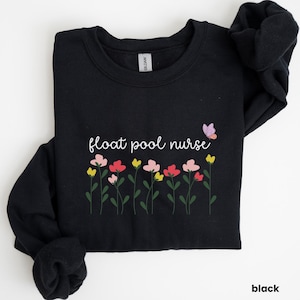 Peut inclure: Sweat-shirt noir avec le texte "float pool nurse" en lettres blanches et un petit papillon violet. Le texte est entouré d'une rangée de fleurs colorées.