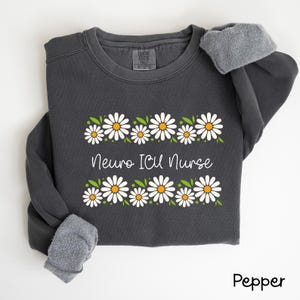 Pode incluir: Uma camisa de moletom cinza escuro com uma borda de margaridas brancas e o texto "Neuro ICU Nurse" em branco. A camisa de moletom tem um forro de l&atilde; cinza.