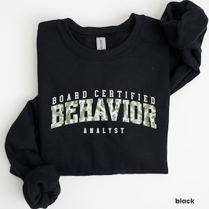 Può includere: Felpa nera con la scritta "BOARD CERTIFIED BEHAVIOR ANALYST" in bianco e verde. La parola "BEHAVIOR" presenta un motivo a felce verde e bianco. La felpa è del marchio Gildan.