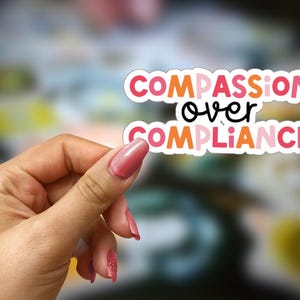 Könnte beinhalten: Ein Aufkleber mit dem Text "COMPASSION over COMPLIANCE" in rosa, orange und weißen Buchstaben auf weißem Hintergrund.