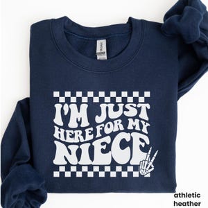May include: A navy blue sweatshirt with a white graphic that says "I'm Just Here For My Niece" with a checkered border and a skeleton hand giving the peace sign.