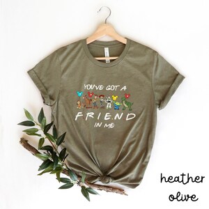 May include: A green t-shirt with the text "You've got a friend in me" and a line of cartoon characters from the movie Toy Story. The characters include Woody, Buzz Lightyear, Jessie, Rex, and Hamm. The shirt is a heather olive color.