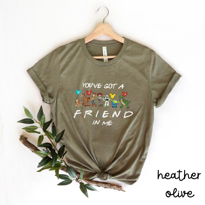 May include: A green t-shirt with the text "You've got a friend in me" and a line of cartoon characters from the movie Toy Story. The characters include Woody, Buzz Lightyear, Jessie, Rex, and Hamm. The shirt is a heather olive color.