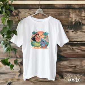 May include: White t-shirt featuring a colorful graphic of Lilo and Stitch in a tropical setting. The design includes palm trees, a sunset, and the characters in Hawaiian attire. The word "white" is printed at the bottom right.