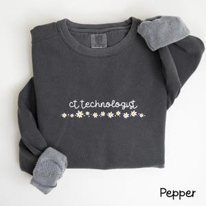 Op de afbeelding: Een donkergrijs sweatshirt met de tekst "ct technologist" in witte letters en een rij witte madeliefjes eronder. Het sweatshirt heeft een fleecevoering.
