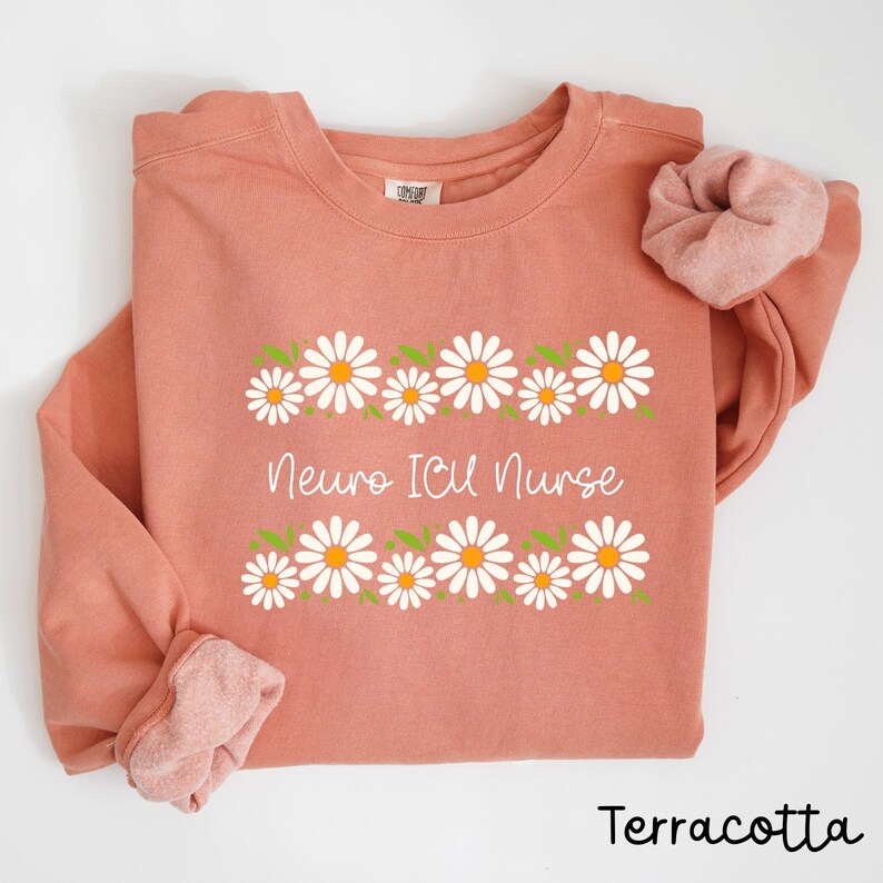 Pode incluir: Camisola de cor terracota com uma borda de margaridas brancas e o texto "Neuro ICU Nurse" em branco.