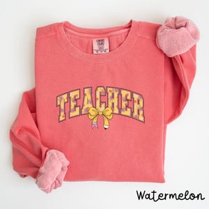 May include: Watermelon coloured sweatshirt with the word "TEACHER" in a yellow arch with a floral pattern and a yellow bow with a pencil inside.