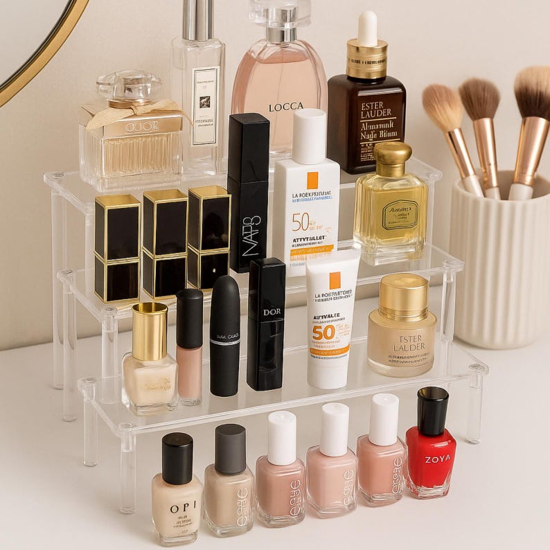 May include: A clear, tiered acrylic organizer displaying various beauty products. Includes perfumes, lipsticks, nail polish, sunscreen, and makeup brushes. The products are in various colors, including gold, black, and pink.