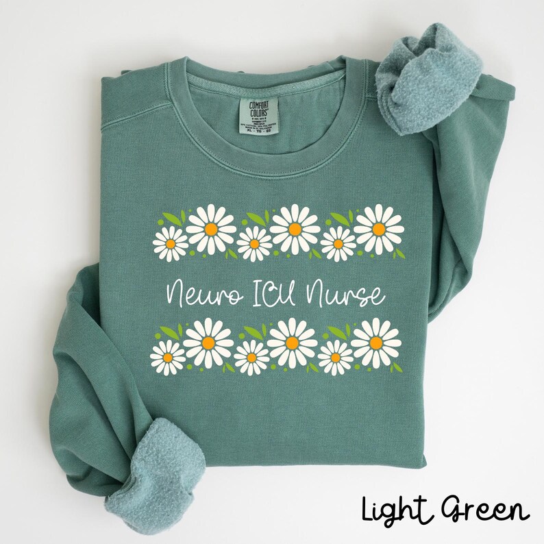 Pode incluir: Camisola verde claro com uma borda de margaridas brancas e o texto "Neuro ICU Nurse" em branco.