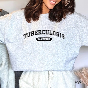 Può includere: Una felpa grigia con la scritta "Tuberculosis Warrior" in lettere nere.