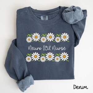 Pode incluir: Uma camisa de frio azul escura com uma borda de margaridas brancas e o texto "Neuro ICU Nurse" em branco. A camisa de frio &eacute; feita de um material macio e confort&aacute;vel.