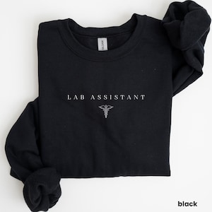 Könnte beinhalten: Schwarzes Sweatshirt mit dem weißen Text "LAB ASSISTANT" und einem weißen Hermesstab-Symbol. Das Kleidungsstück besteht aus einem weichen Material und eignet sich für den Freizeitgebrauch. Das Wort "black" ist unten rechts aufgedruckt.
