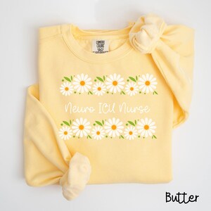 Pode incluir: Uma camisa de moletom amarela com uma borda de margaridas e o texto "Neuro ICU Nurse" impresso na frente. A camisa de moletom &eacute; feita de um material macio e confort&aacute;vel e &eacute; perfeita para o uso di&aacute;rio.
