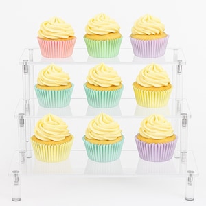 May include: Nine cupcakes with yellow frosting in pastel-colored paper cups are arranged on a clear, three-tiered display stand. The cupcakes are in pink, green, yellow, and purple cups, creating a colorful presentation.