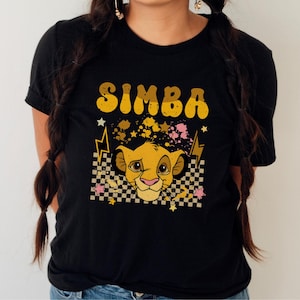 Puede incluir: Camiseta negra con un diseño de Simba. El diseño incluye la palabra "SIMBA" en amarillo, un león de dibujos animados, estrellas y rayos. El león está sobre un fondo a cuadros.