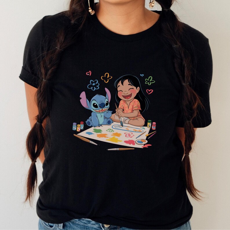 May include: Black t-shirt featuring a colorful illustration of Lilo and Stitch painting together. The image shows Lilo and Stitch sitting on a white sheet of paper with paint supplies. The design includes colorful paint splatters, flowers, and hearts.