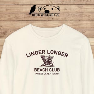 May include: Cream-colored sweatshirt with the text "Linger Longer Beach Club Priest Lake - Idaho" in brown. The design includes a bear relaxing in a beach chair. The brand logo "Bird & Bear Co." is at the top.
