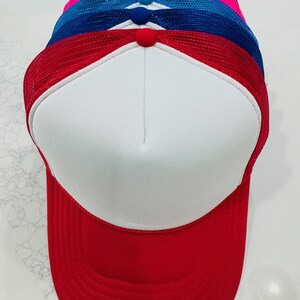 May include: A stack of trucker hats with white front panels and colored mesh backs. The hats are red, blue, pink, and black. Each hat has a matching colored button on top.