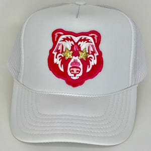 May include: White trucker hat with a pink bear wearing gold glitter star sunglasses. The bear is embroidered on the front of the hat.