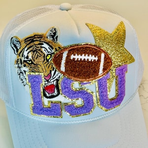 May include: White trucker hat with a purple and gold LSU logo, a brown football, a tiger, and a gold glitter star.