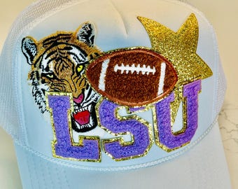 May include: White trucker hat with a purple and gold LSU logo, a brown football, a tiger, and a gold glitter star.
