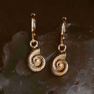 May include: A pair of gold-toned hoop earrings featuring small, dangling charms shaped like spiral ammonite fossils. The earrings have a polished metallic finish and a hinged closure mechanism, showcasing a detailed, textured shell pattern on each charm.