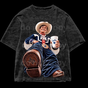 Op de afbeelding: Een antracietgrijs acid-wash T-shirt met een afbeelding van een cowboy in een witte hoed, blauw jasje met franjes en een spijkerbroek. De cowboy houdt een witte beker vast met het opschrift Dallas en een Texas-ster, de zool toont Texas.