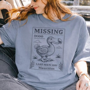 Op de afbeelding: Lichtblauw T-shirt met een afbeelding van een Dodo-vogel en de tekst "MISSING" en "LAST SEEN 1681 Mauritius". Het shirt wordt gedragen met een bruine pet en zonnebril.