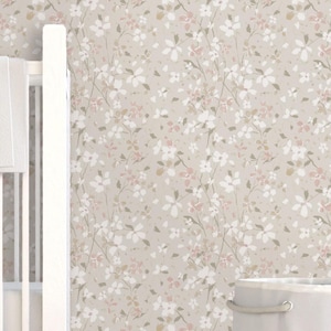 May include: A white cot with a white blanket draped over the side. The cot is in front of floral wallpaper with white, pink, and green flowers on a beige background. A white laundry basket sits on a white rug.