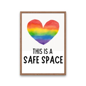 May include: A framed art print with a rainbow heart design. The text "THIS IS A SAFE SPACE" is displayed below the heart in bold black letters. The artwork is set against a white background, within a wooden frame.