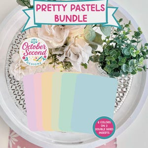 May include: A bundle of pastel-colored inserts, including pink, yellow, green, and blue, arranged on a white tray with floral decorations. The text "Pretty Pastels Bundle" is displayed above the inserts, with a logo that says "October Second Designs". The inserts are double-sided.