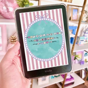 Puede incluir: Un lector de libros electrónicos Kindle con una funda transparente muestra un gráfico colorido. El gráfico presenta el texto "STAYLOR'S KINDLE" en un círculo con elementos decorativos sobre un fondo a rayas. El lector de libros electrónicos se sostiene en una mano.