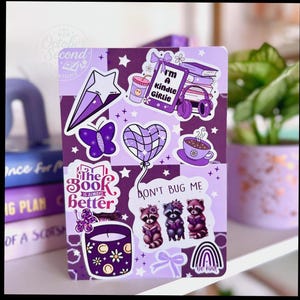 May include: A purple and white checkered notebook covered in stickers. Stickers include a star, butterfly, heart-shaped balloon, coffee cup, raccoons, and the text "I'm a Kindle Girlie" and "The Book is Always Better".