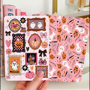 May include: Pink Halloween-themed sticker sheet with various framed illustrations. The designs include ghosts, pumpkins, bows, and a haunted house. The sheet is held by a person's hand.