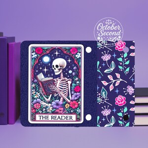 May include: A tarot card featuring a skeleton reading a book, surrounded by flowers and stars, with the text "THE READER." The card has a navy blue background and is part of a notebook with a floral pattern and the text "October Second Designs."