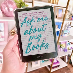 Puede incluir: Un lector electrónico Kindle con una funda transparente muestra el texto "Ask me about my Books" en escritura turquesa. La pantalla también muestra un diseño de tarjeta de biblioteca vintage. El Kindle se sostiene frente a un fondo borroso.
