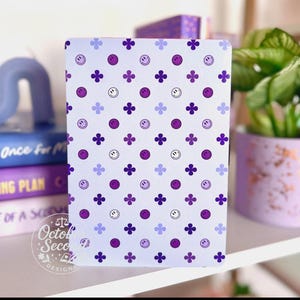 May include: A light purple notebook with a repeating pattern of purple smiley faces, four-leaf clovers, and small purple dots. The notebook is on a white shelf with books and a potted plant.