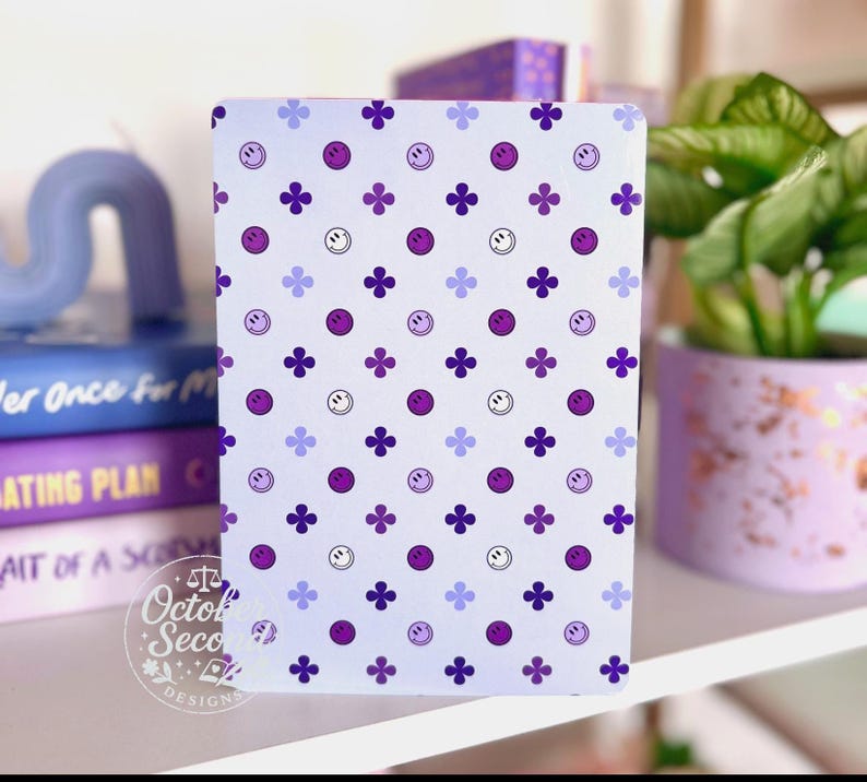 May include: A light purple notebook with a repeating pattern of purple smiley faces, four-leaf clovers, and small purple dots. The notebook is on a white shelf with books and a potted plant.