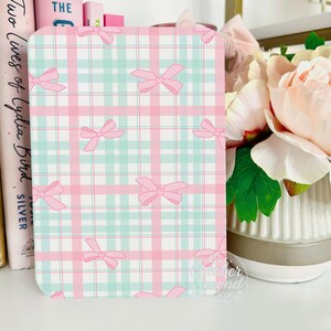 A pink and green plaid notebook with bows on a white surface, next to a vase with pink flowers.