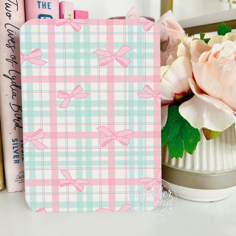 A pink and green plaid notebook with bows on a white surface, next to a vase with pink flowers.