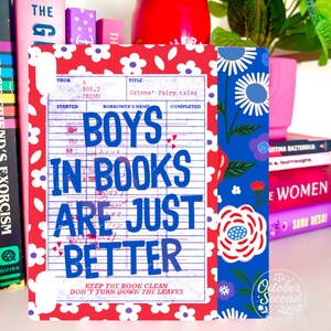 May include: A decorative book with a red and white floral pattern and the text "BOYS IN BOOKS ARE JUST BETTER" in blue. The book has a vintage library card design and is surrounded by other books.