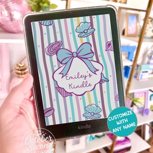 以下が含まれることがあります： 透明な保護ケースに入ったKindle電子書籍リーダー。画面には水色、紫、白の縦縞模様のスキンが貼られている。デザインには紫色の貝殻のイラスト、大きな水色のリボン、そして「Emiley's Kindle」というテキストが含まれている。